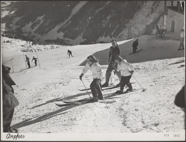 Gruppe von Menschen, die eine schneebedeckte Piste hinunterskifahren, Ski-Stöcke haltend, mit Hügeln und einem Gebäude im Hintergrund und Text unten.