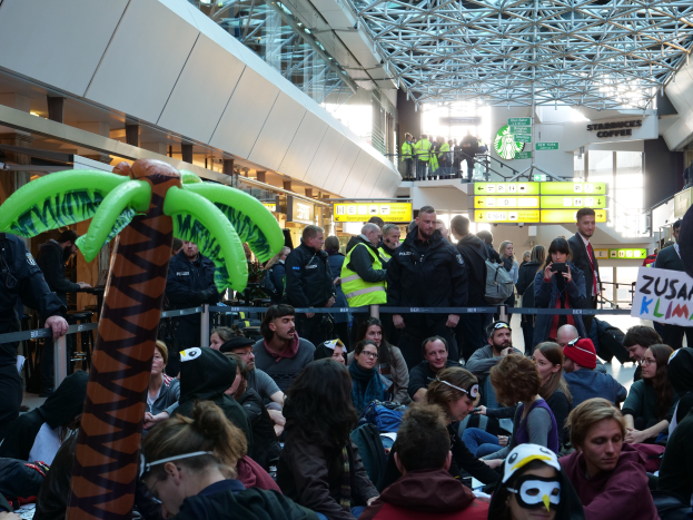 Eine große Gruppe von Menschen sitzt in einem Flughafen, einige tragen Mützen und Schutzbrillen, andere halten Schilder, mit einem Geländer im Vordergrund und Texttafeln im Hintergrund, unter einer Decke, die mit Stangen und Lichtern geschmückt ist.