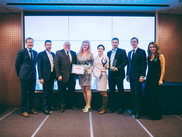 Gruppe von strahlenden Menschen mit Zertifikaten vor einem Bildschirm, mit einem Tisch und einem Wandobjekt im Hintergrund, Gewinner der International Business Awards 2018.