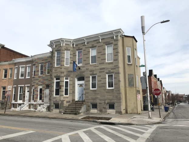 Eine Reihe von Häusern an einer Straßenecke in Philadelphia, mit Fenstern, Türen, Geländern, Laternenmasten, Schildern und Fahrzeugen, sowie Bäumen und einem bewölkten Himmel im Hintergrund.