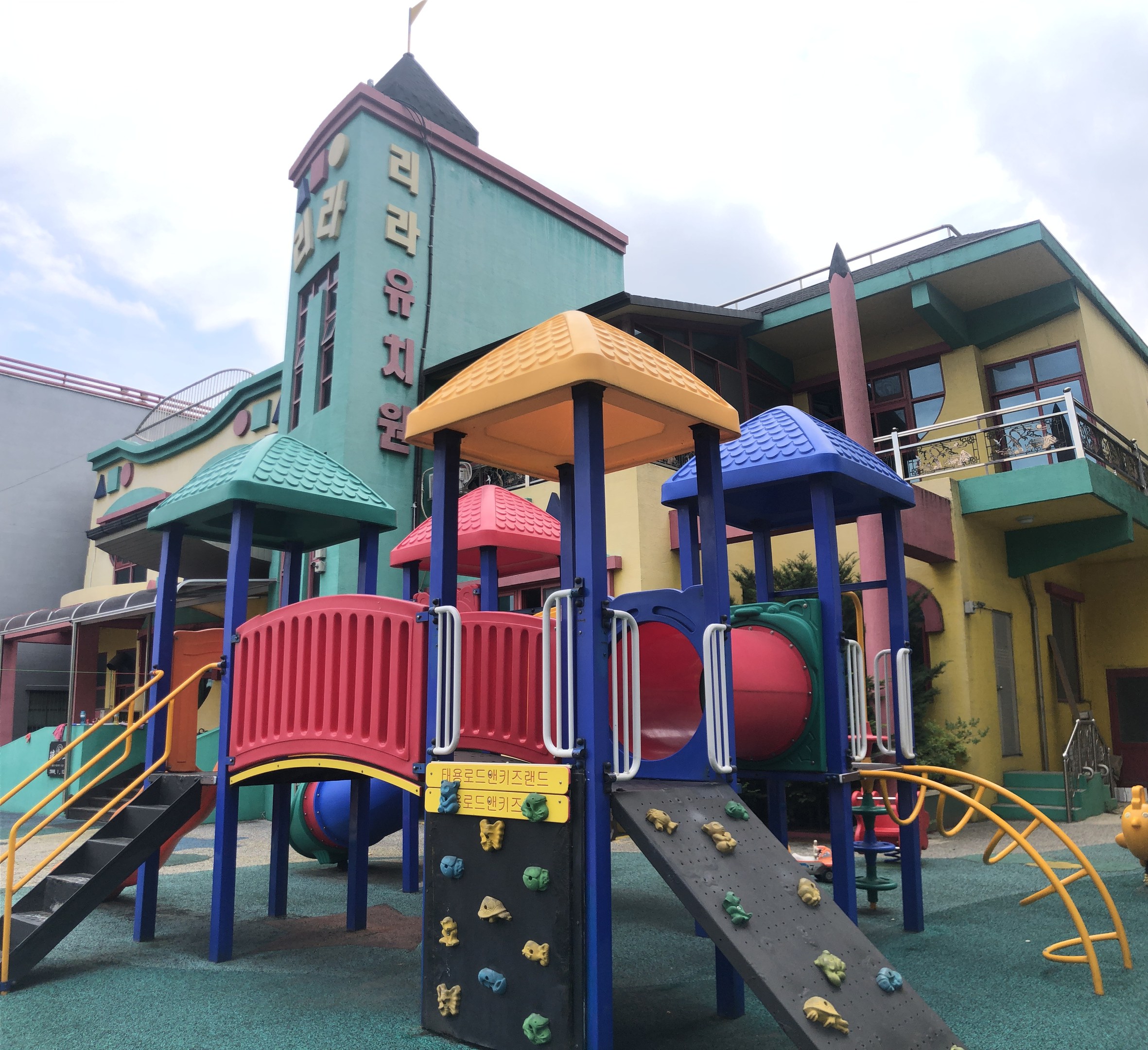 Buntes Kinderspielplatzgerät vor einem Gebäude mit Fenstern, umgeben von Bäumen und einem bewölkten Himmel.