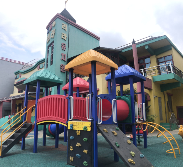 Buntes Kinderspielplatzgerät vor einem Gebäude mit Fenstern, umgeben von Bäumen und einem bewölkten Himmel.