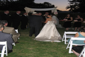 Eine Gruppe von Menschen bei einer Hochzeit, wobei die Frauen auf der rechten Seite Blumenbouquets halten, während andere auf Stühlen sitzen, umgeben von Bäumen.
