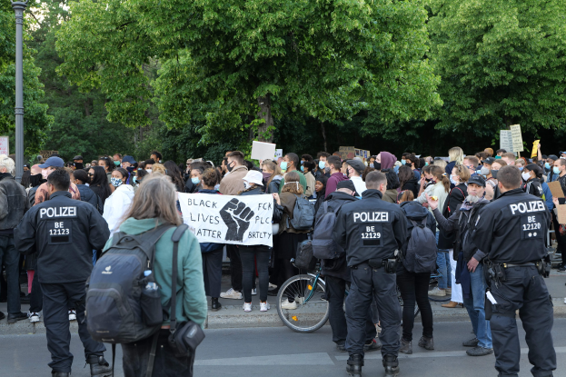 Eine große Gruppe von Menschen steht an der Straße, einige halten Schilder, mit einem Fahrrad im Vordergrund und Bäumen und einem Mast im Hintergrund, bei einer Black-Lives-Matter-Demonstration in Berlin.