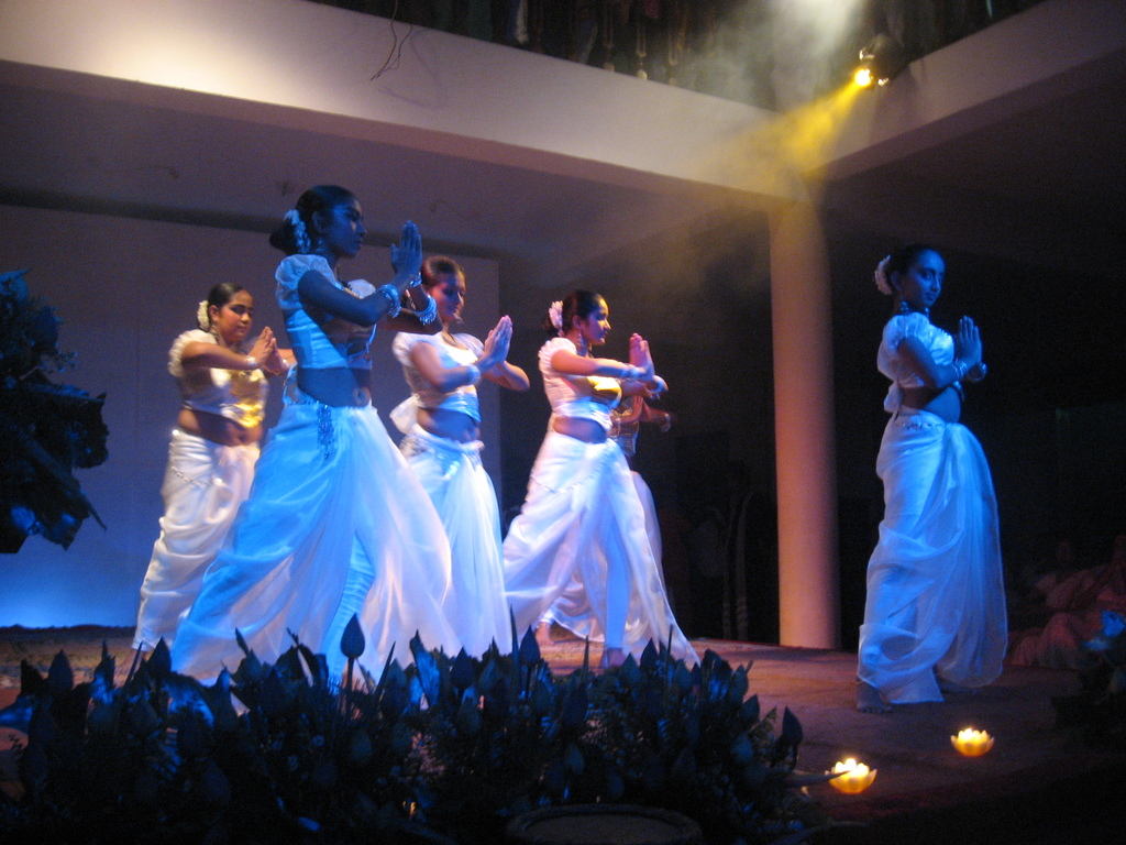 Eine Gruppe von Menschen, die auf einer Bühne mit Pflanzen davor und einer anderen Gruppe, die zusieht, tanzt.