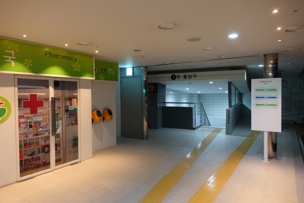 Apothekeneingang mit einem "Apotheke"-Schild auf der linken Seite, einer Tafel mit Text auf der rechten Seite, Hintergrundobjekten und beleuchtetem Dach.