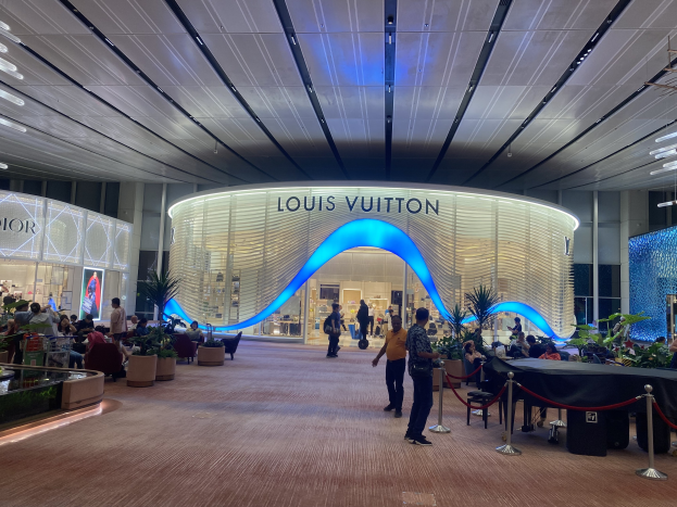 Louis Vuitton Store am Hong Kong International Airport mit sitzenden und stehenden Menschen, umgeben von Absperrpollern, Display-Elementen und Deckenleuchten.