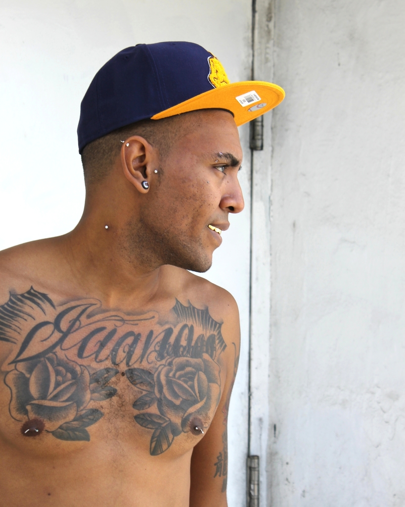 Eine Person mit einer Mütze mit einem Aufkleber mit einem Barcode und Bildern, mit sichtbaren Tattoos auf dem Körper, die vor einer Tür und einer Wand steht.