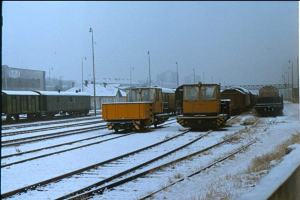 Eine Gruppe von Zügen auf schneebedeckten Schienen mit Pfählen, Gebäuden und einer Brücke im Hintergrund, unter einem sichtbaren Himmel, mit Text unten.