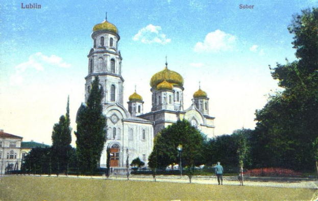 Old photograph of the Church of the Intercession on the Nerl in Lublin, Russia, featuring its golden domes, surrounded by trees, street poles, lights, and pedestrians under a cloudy sky with text at the top.