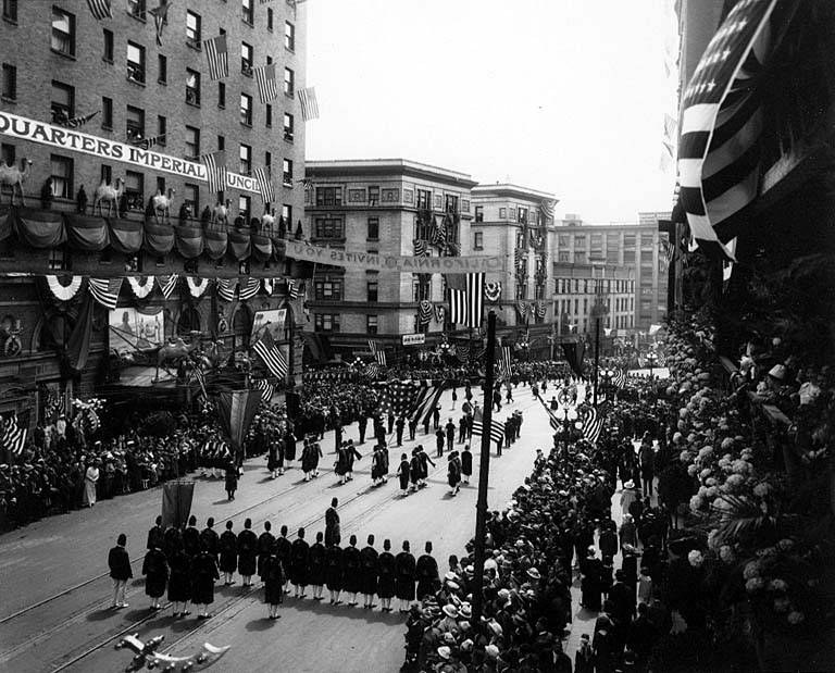 A large crowd of people walks down a New York City street during a Fourth of July parade, many wearing caps and holding flags, with others standing along the sides.