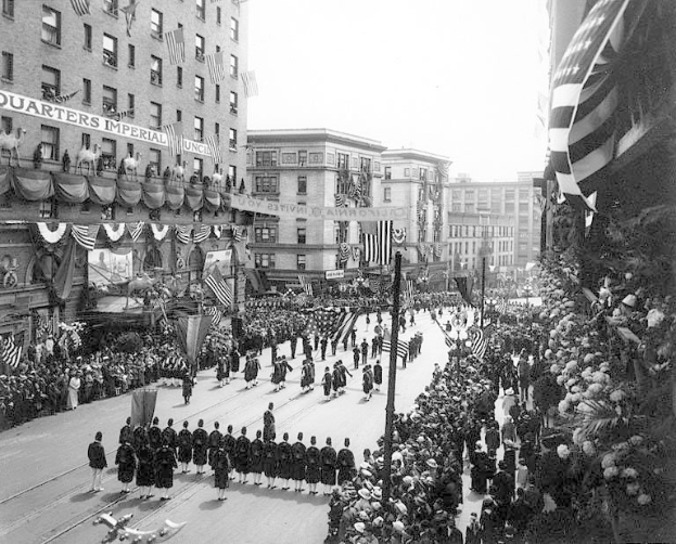 A large crowd of people walks down a New York City street during a Fourth of July parade, many wearing caps and holding flags, with others standing along the sides.