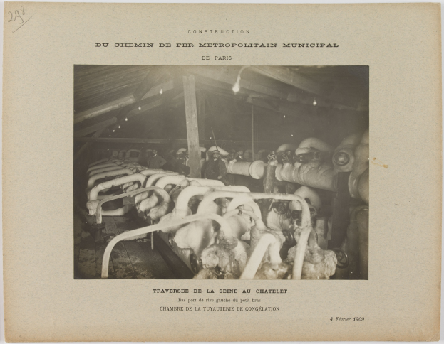 Schwarz-weiß-Foto von Arbeitern in einer Fabrik umgeben von Rohren und beleuchtet von Deckenlampen, mit Text unten.