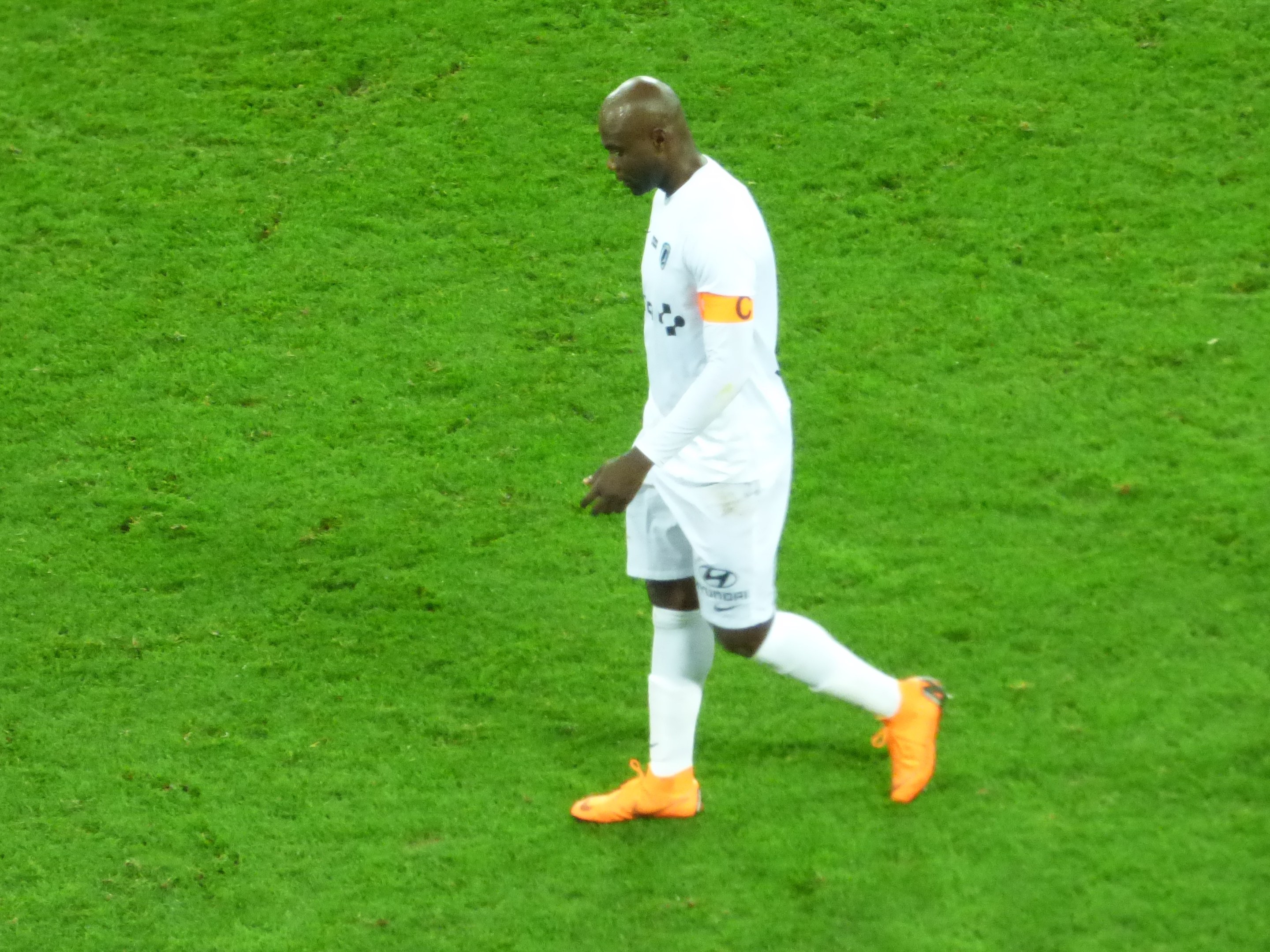 A soccer player in a white t-shirt, white shorts, and orange shoes walks across a lush green field.