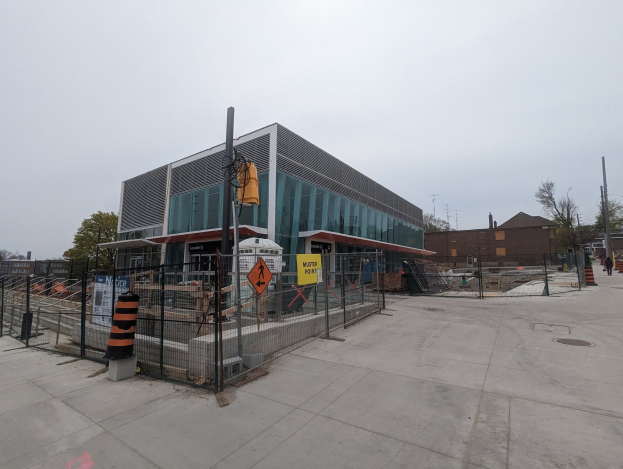 Large multi-story school building with numerous windows, surrounded by a metal fence, trees, signboards, utility poles, pedestrians, and vehicles under a cloudy sky.