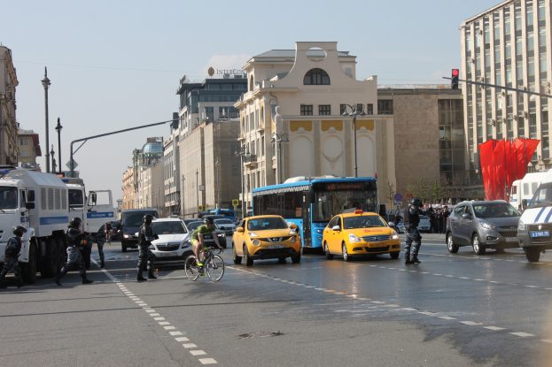 A busy city street with cars, trucks, bicycles, pedestrians in helmets holding guns, light poles, traffic signals, buildings with windows, and a cloudy sky.