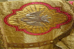 Nahaufnahme eines Priestergewandes mit aufwendiger Gold- und Rotstickerei vor einem neutralen Wandhintergrund.