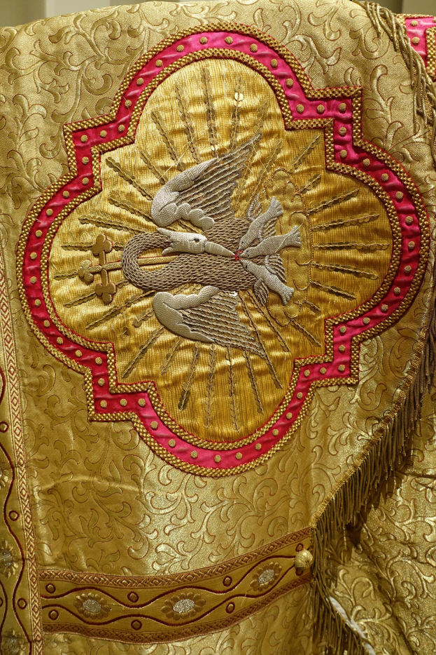 Nahaufnahme eines Priestergewandes mit aufwendiger Gold- und Rotstickerei vor einem neutralen Wandhintergrund.