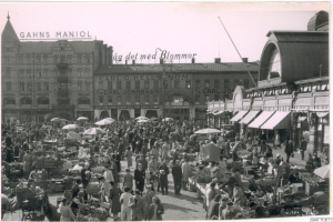 Schwarzes und weißes Foto eines belebten Berliner Markts mit Menschen, Gemüsewagen und Gebäuden im Hintergrund.