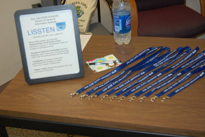 Ein Tisch mit ID-Kartenhaltern, ein Photo-Rahmen, eine Karte, ein Stift, eine Wasserflasche und ein Stuhl mit einer Tasche darauf.