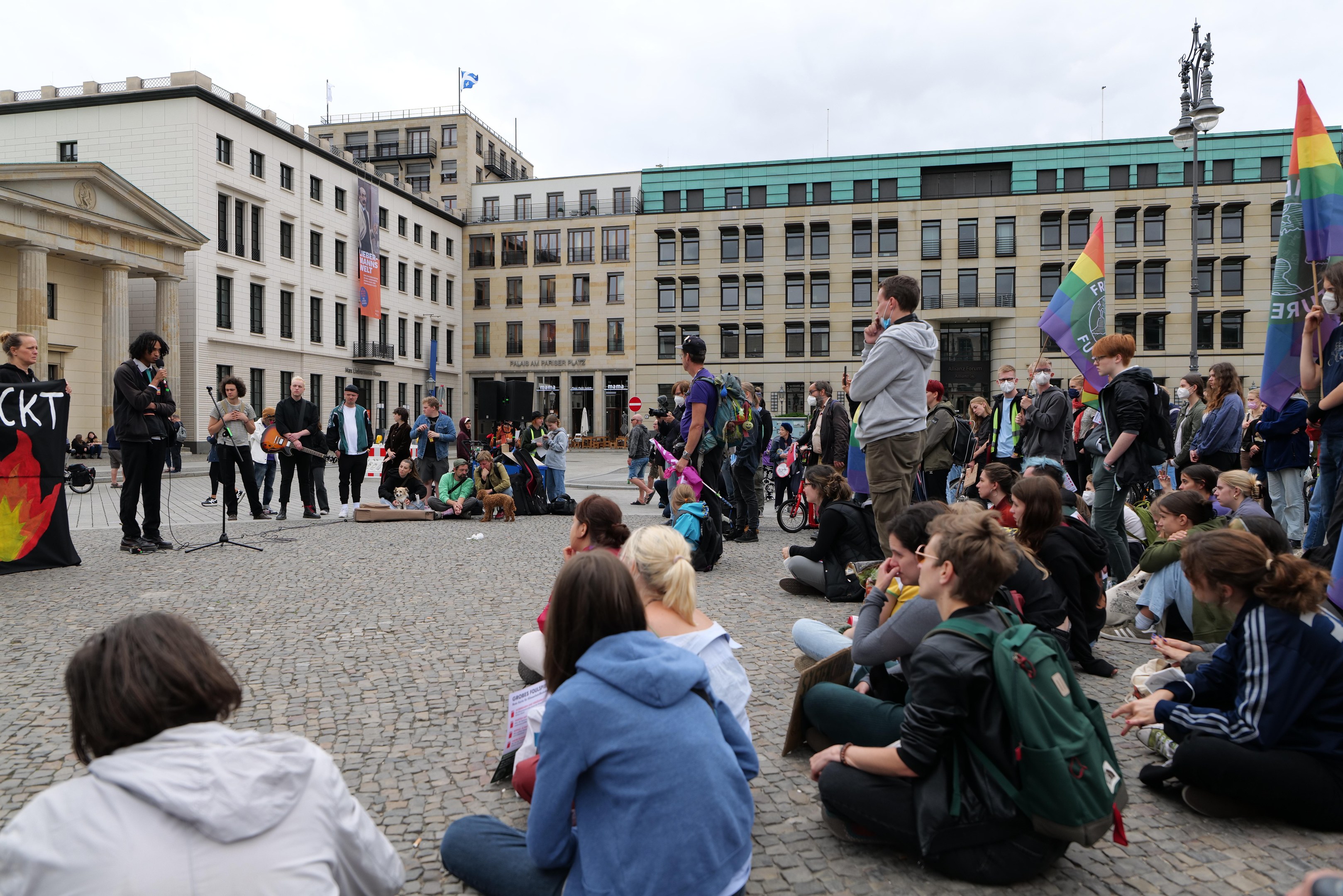 Eine Gruppe von Menschen, die auf dem Boden vor einer Menge mit Fahnen und Spruchbändern sitzen, mit einer Person, die in ein Mikrofon spricht, einer Statue und Gebäuden im Hintergrund während einer anti-schwulen Demonstration in Berlin, Deutschland.