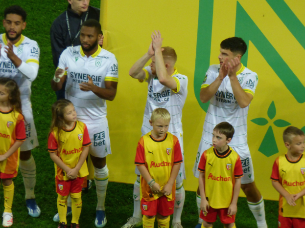 A group of soccer players in uniforms clapping in celebration on a Ligue 1 field, with a "Ligue 1" sign visible in the background.