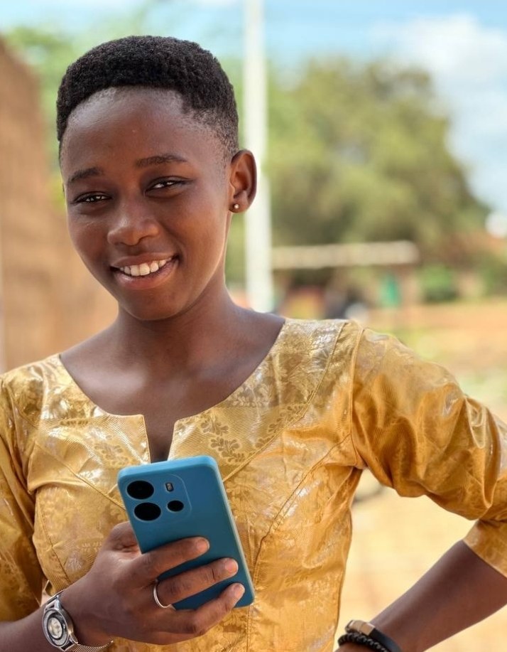 Ein junges Mädchen in einem gelben Kleid hält ein blaues Handy und strahlt vor einem Hintergrund aus Wand, Pfahl, Bäumen und Himmel.