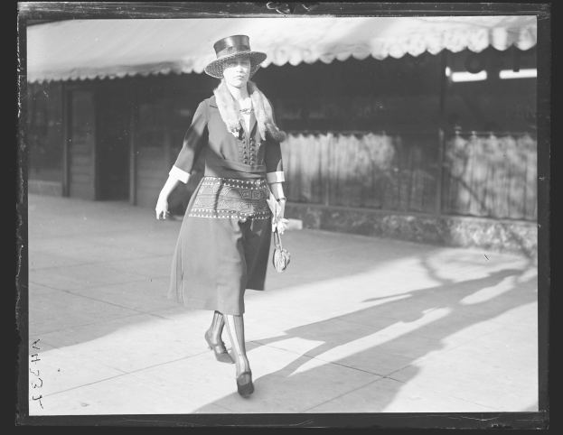 Frau in einem Kleid und Hut, die die Straße entlang geht und eine Handtasche hält, trägt hohe Absätze, in Schwarz-Weiß.