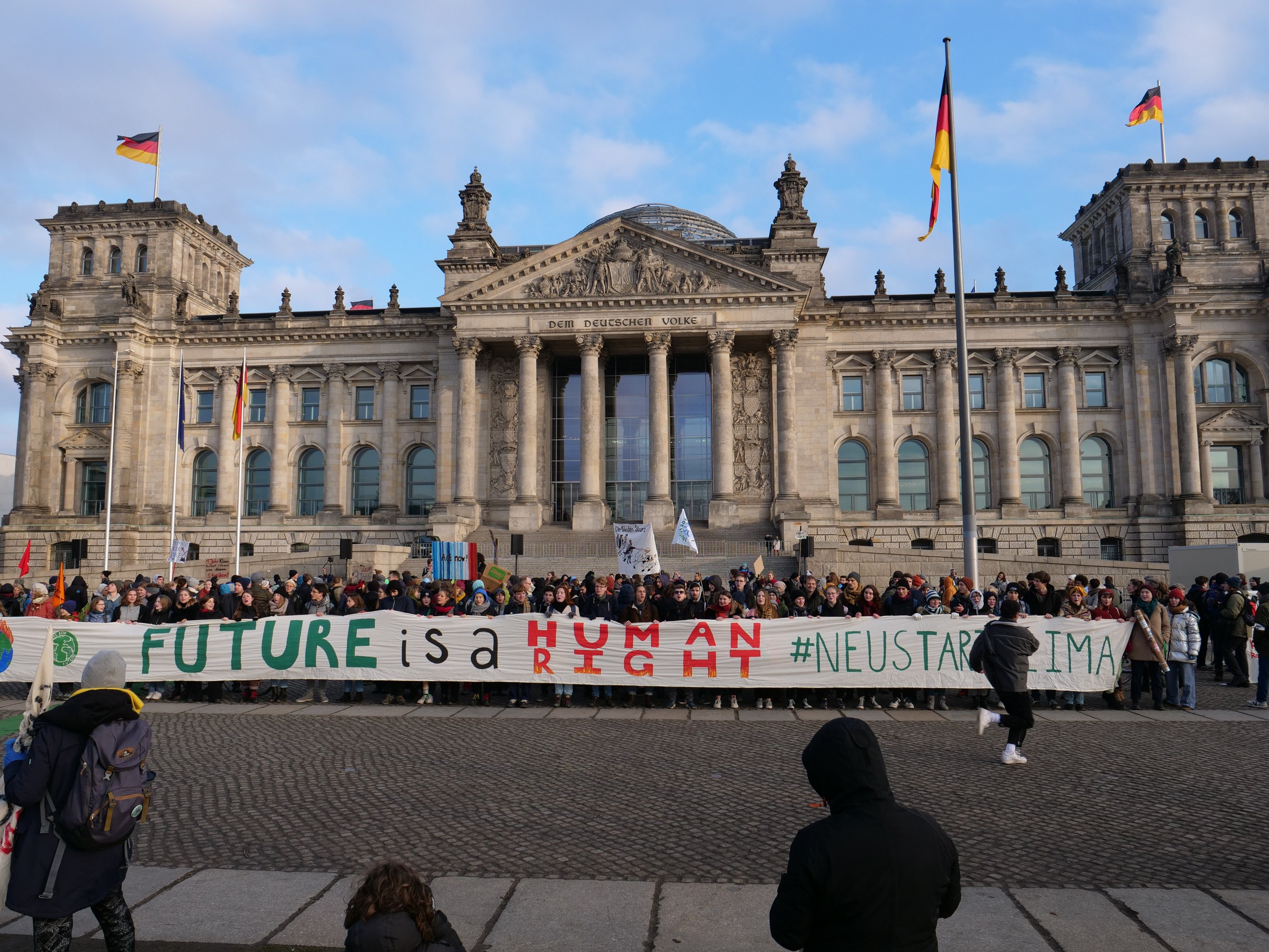 Group of people standing in front of the Reichstag building in Berlin, holding a banner with the text "Future is a Human Neustar ima," with the building's architectural details visible and flags surrounding it under a cloudy sky.