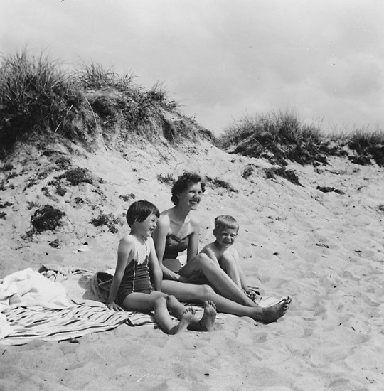 Schwarzes und weißes Foto einer Frau in einem Bikini und zwei Kindern in Badebekleidung, die auf einer Decke am Strand sitzen, mit Sand, Gras und Himmel im Hintergrund.