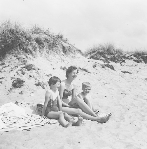 Schwarzes und weißes Foto einer Frau in einem Bikini und zwei Kindern in Badebekleidung, die auf einer Decke am Strand sitzen, mit Sand, Gras und Himmel im Hintergrund.