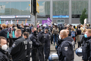 Eine große Gruppe von Menschen steht vor einem Gebäude, einige halten Schilder und tragen Helme, mit einem Mast mit einer Schildertafel im Vordergrund und einem Baum im Hintergrund, die zu protestieren scheinen.