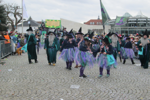 Eine Gruppe von Menschen in Hexenkostümen, die eine Kopfsteinpflasterstraße entlanggehen. Sie tragen schwarze und lila Kleider und Hüte und halten einige Fahnen. Auf der linken Seite der Straße befindet sich ein Geländer, und im Hintergrund sind Gebäude, Bäume und ein klarer blauer Himmel zu sehen.