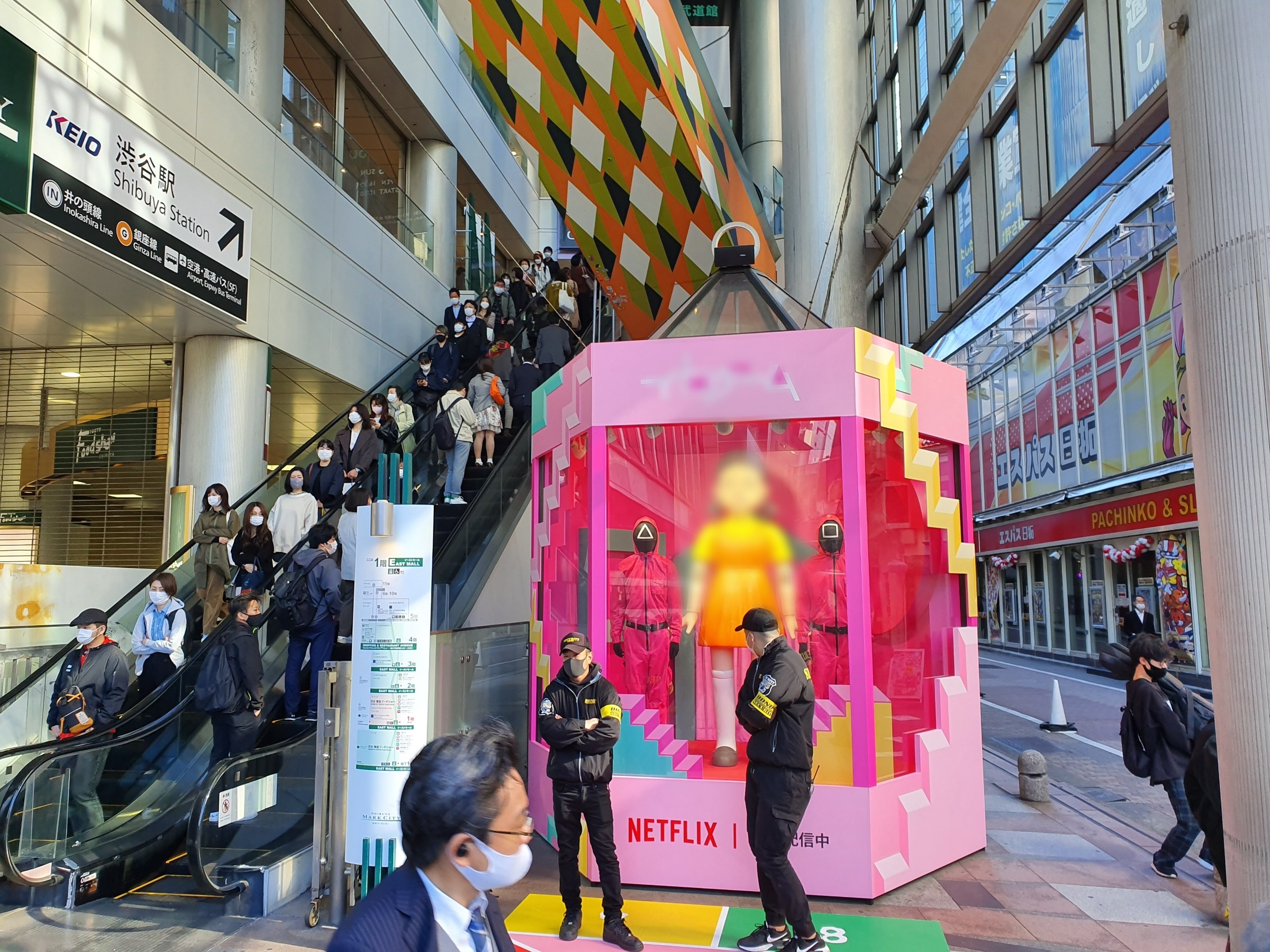 Eine Gruppe von Menschen steht vor einem Gebäude mit Glaswänden, Säulen und Texttafeln, einige tragen Masken, mit einer Rolltreppe im Blick; eine pinkfarbene Box in der Mitte zeigt Schaufensterpuppen und trägt die Aufschrift "Netflix's new Netflix-Boot in Tokyo."