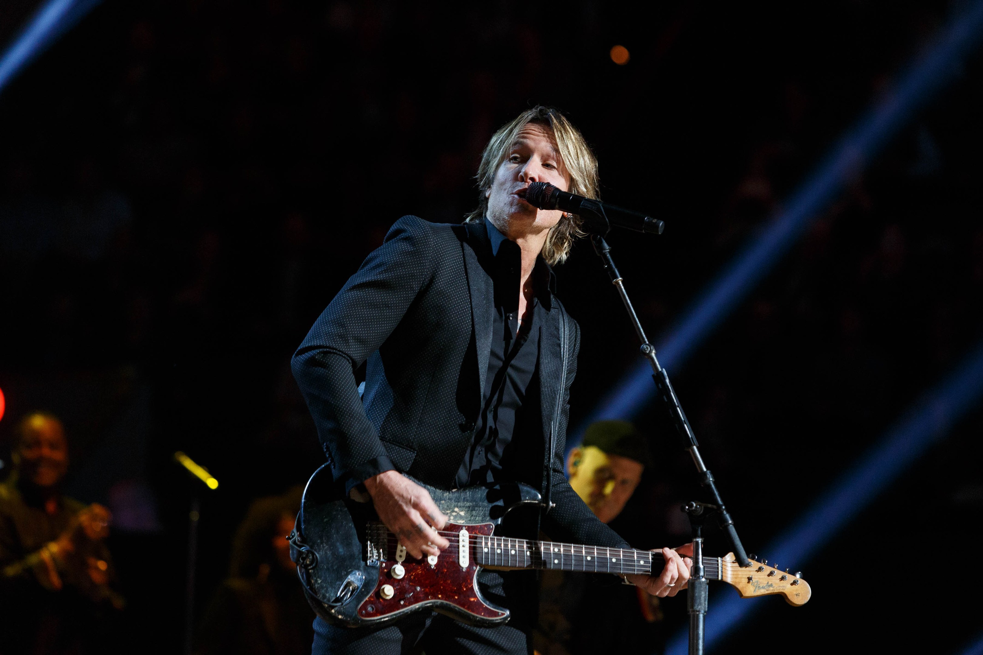 Keith Urban performing on stage at the Joint at Hard Rock Hotel & Casino in Las Vegas, playing guitar with a microphone and band members visible in a dimly lit setting.