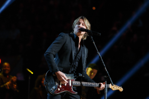 Keith Urban performing on stage at the Joint at Hard Rock Hotel & Casino in Las Vegas, playing guitar with a microphone and band members visible in a dimly lit setting.