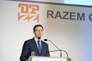 A suited man stands at a podium with a glass of water and two microphones, speaking as CEO of Razem Global, with the company banner behind him.