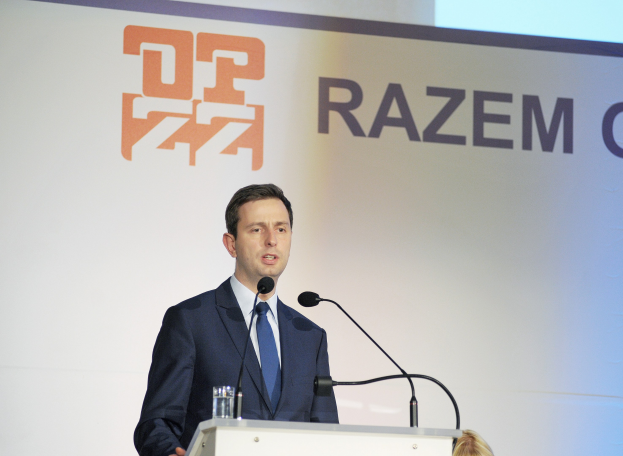 A suited man stands at a podium with a glass of water and two microphones, speaking as CEO of Razem Global, with the company banner behind him.