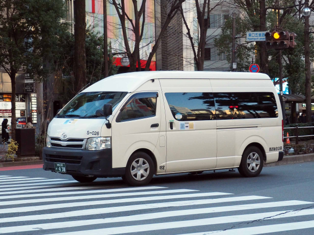 Ein weißer Lieferwagen fährt eine Straße mit hohen Gebäuden, Bäumen, Pfählen, Verkehrsampeln, Schildern, Pflanzen und Menschen entlang.