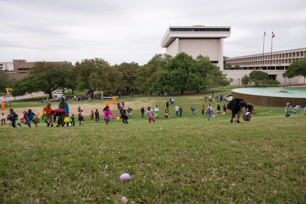 Gruppe von Menschen, die mit Ostereiern in einem grasbewachsenen Feld mit Bäumen, Gebäuden, Fahnen, einem Brunnen und einem bewölkten Himmel im Hintergrund spielt.