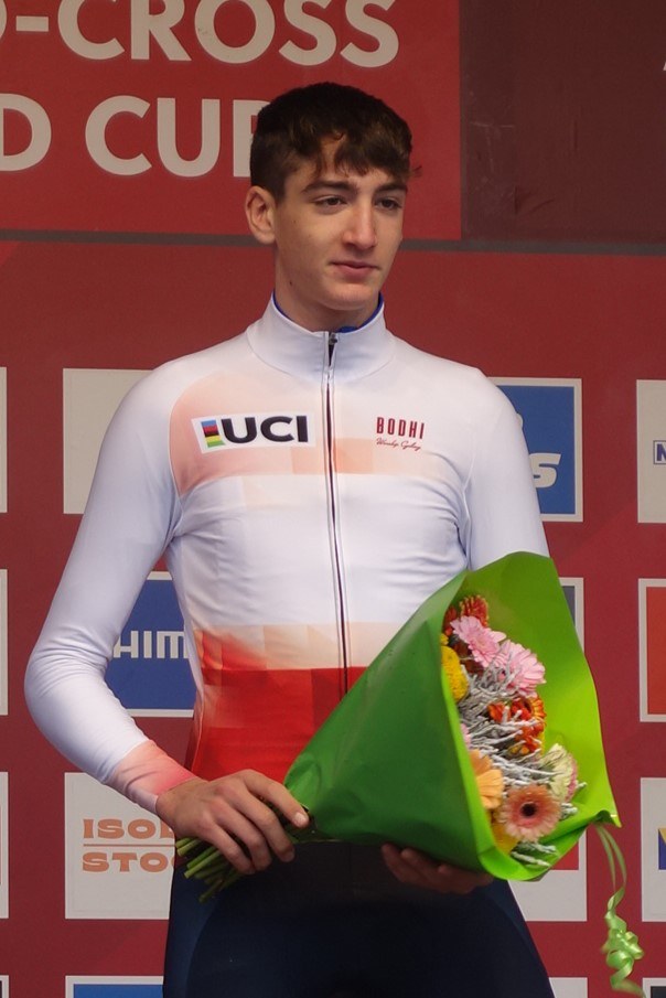 A man stands on a podium holding a bouquet of flowers, celebrating with a confident and proud expression.