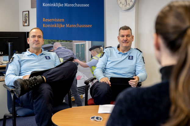Zwei Polizeibeamte sitzen an einem Tisch mit einer Frau, umgeben von Papieren und Gegenständen, mit einem Wandbanner, einer Uhr und Foto Rahmen im Hintergrund und einem weiteren Tisch mit einem Monitor, Tastatur und Maus links.