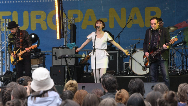 Vier Personen auf der Bühne: eine Frau in einem weißen Kleid singt in der Mitte, flankiert von zwei Männern, die Gitarren spielen, mit einer sichtbaren Menge und Lautsprechern und einer Wand im Hintergrund.