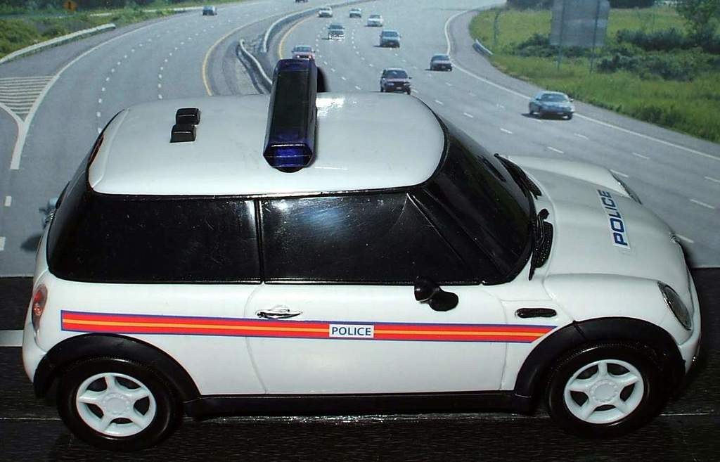 White police car in front of moving traffic on a road lined with grass and trees outside the city.
