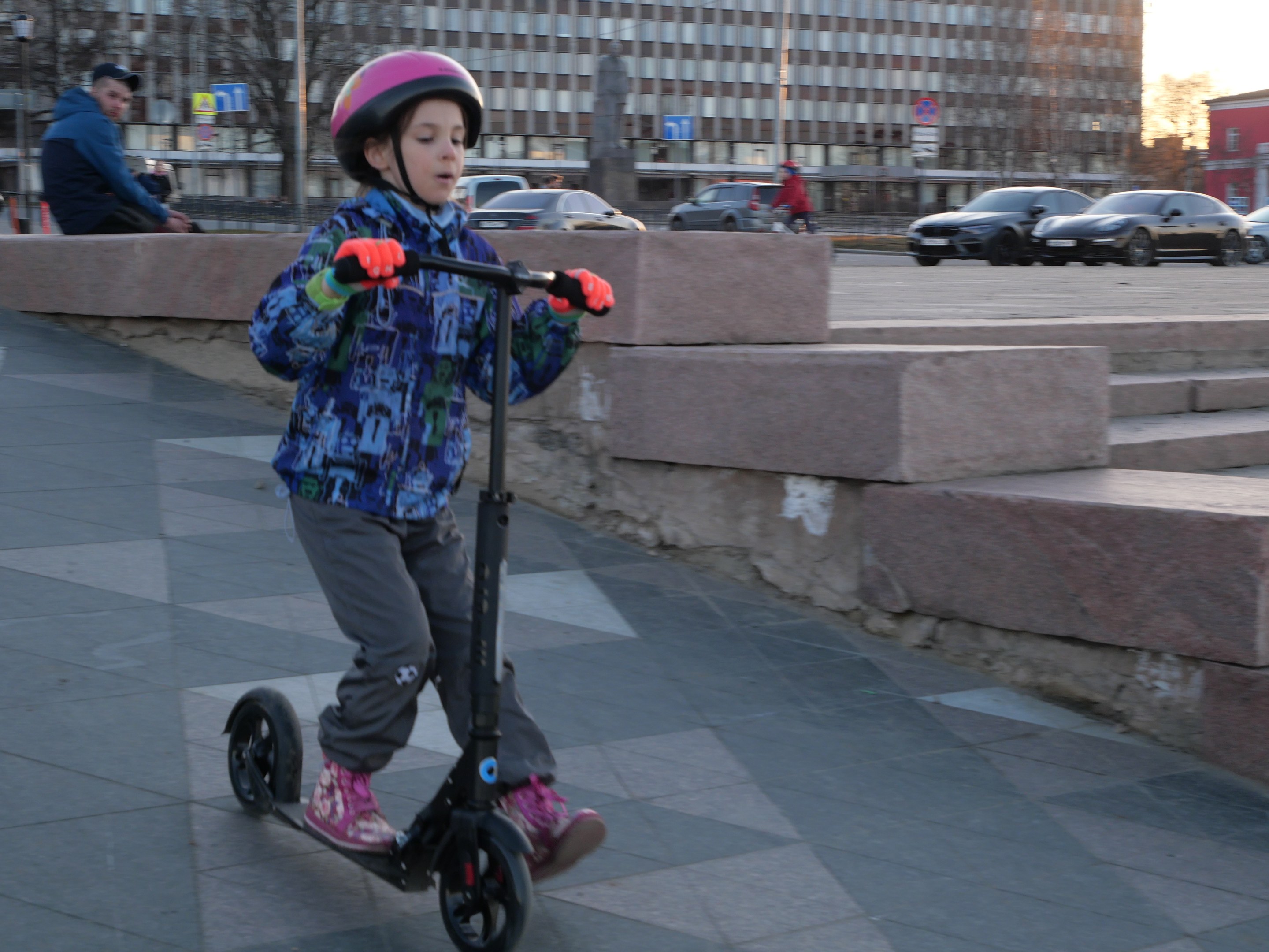 Ein junger Junge mit Helm und Handschuhen fährt einen Roller eine Treppe hinunter, vorbei an Fahrzeugen, Menschen, Bäumen, Pfosten, Brettern, Gebäuden und einem klaren blauen Himmel.