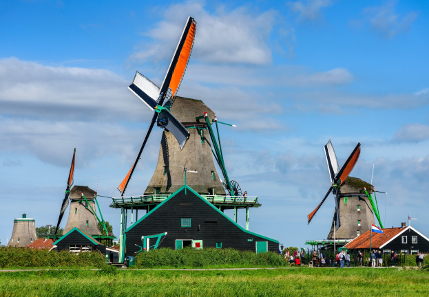 Gruppe von traditionellen Windmühlen in Kinderdijk, Niederlande, mit Menschen in der Nähe, umgeben von grünem Gras, Pflanzen, Bäumen und einem klaren blauen Himmel.