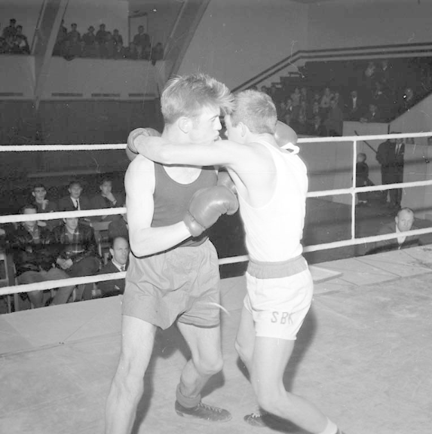 Zwei Boxer kämpfen in einem Boxring, umgeben von sitzenden und stehenden Zuschauern, auf einem Schwarz-Weiß-Bild.