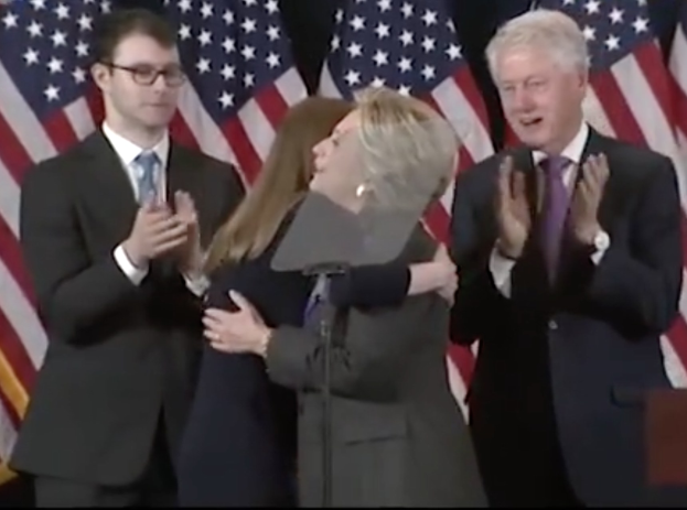 Hillary Clinton und Bill Clinton klatschen vor einem Rednerpult mit Mikrofon, umgeben von Flaggen.