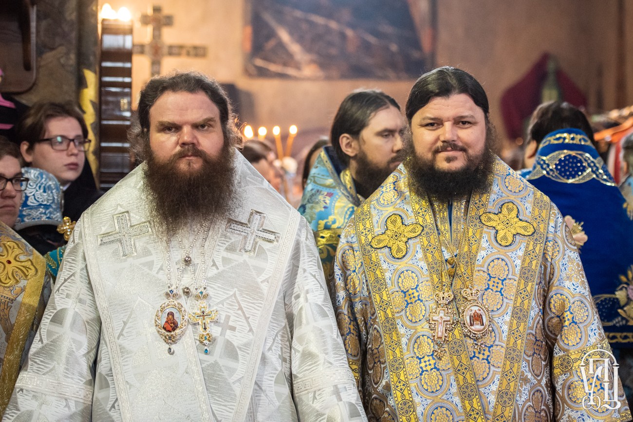 Zwei russische orthodoxe Priester in traditioneller Kleidung stehen nebeneinander in einer Kirche, umgeben von einer Gruppe von Menschen in bunten Kleidern, mit gerahmten Kunstwerken an der Wand und Gegenständen links.