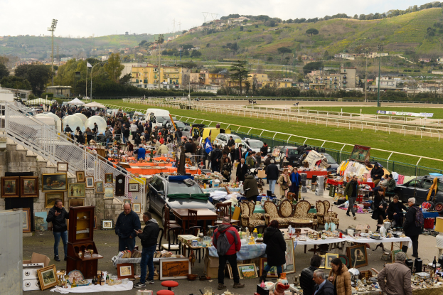 Eine große Gruppe von Menschen auf einem Flohmarkt mit Tischen, auf denen Gegenstände wie Foto Rahmen und Stühle ausgelegt sind, Fahrzeuge in der Nähe geparkt, Geländer, Treppen, Bäume, Gebäude, Laternenpfähle, Hügel und ein bewölkter Himmel im Hintergrund.
